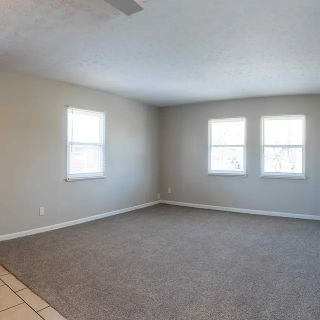A spacious empty living room featuring two windows on the right side and a kitchen area on the left with wooden cabinets.