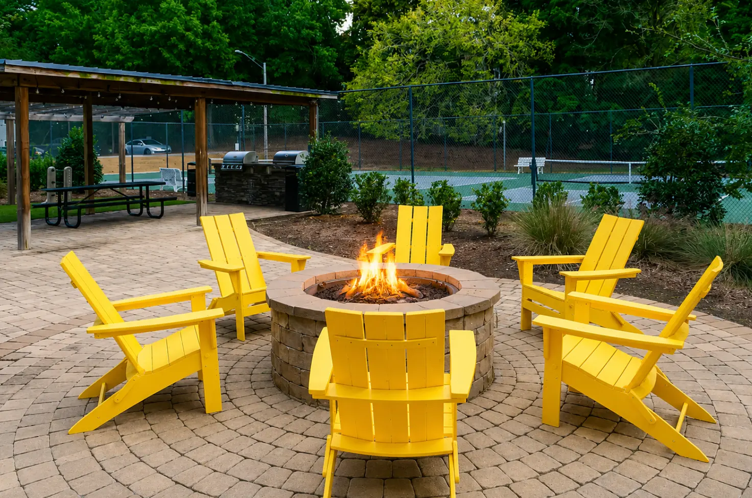 A circular stone fire pit surrounded by bright yellow Adirondack chairs, set in a landscaped area with greenery in the background.