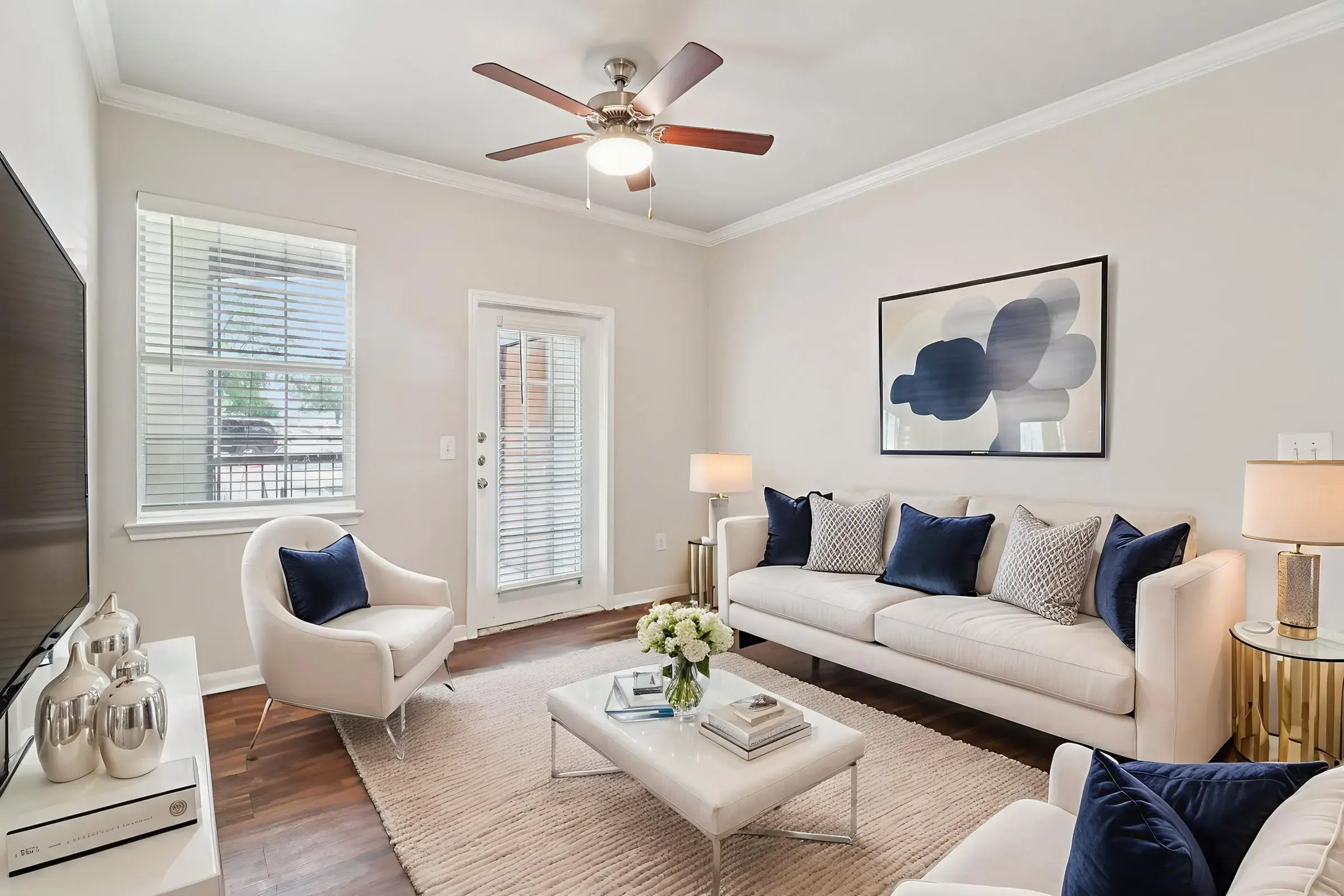 A modern living room featuring a white sofa with blue accent pillows, two white chairs, and a coffee table. A ceiling fan is visible above, and there are large windows allowing natural light in. The walls are painted light colors, with a simple artwork hanging above the sofa.