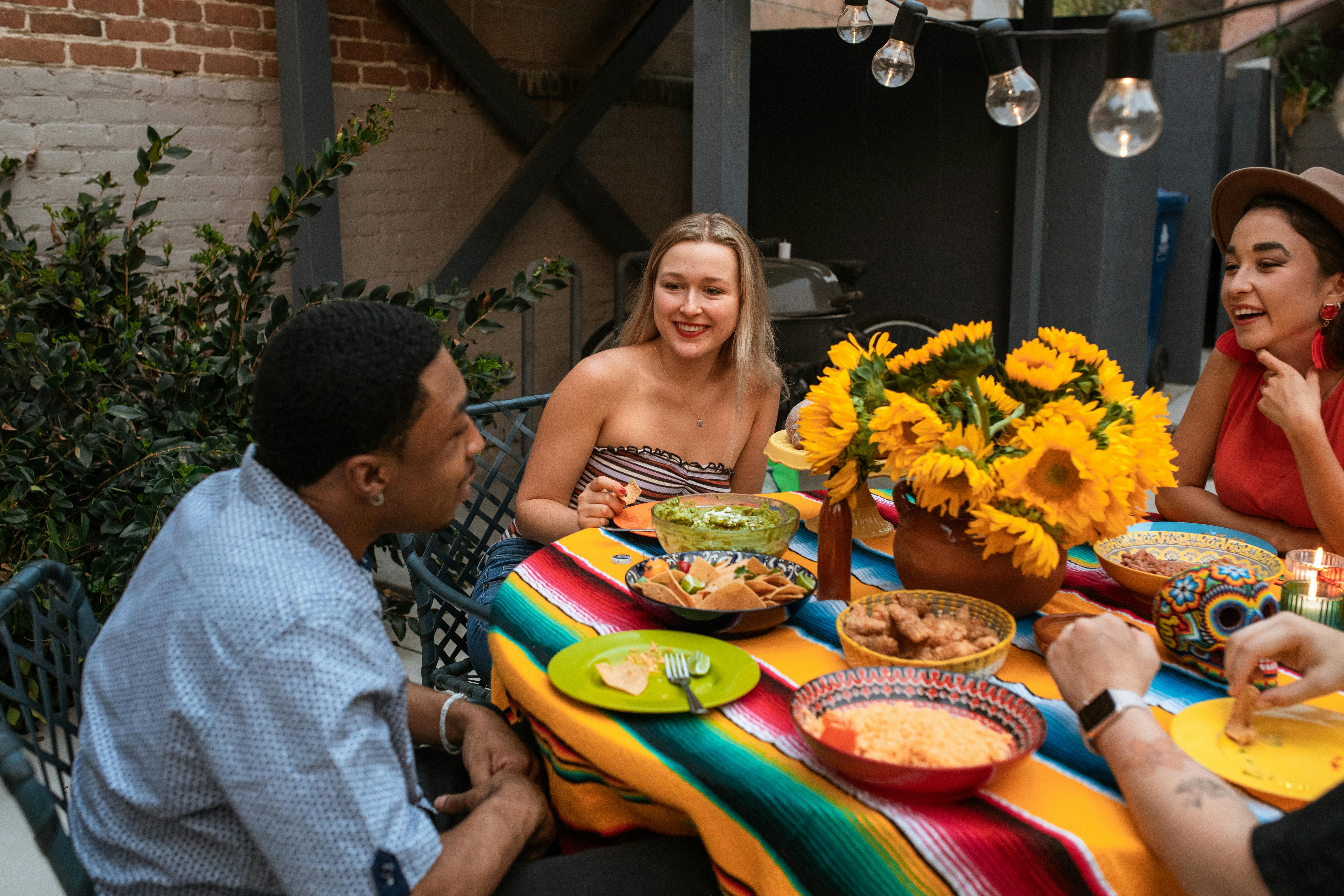 A group of friends enjoying a lively dinner outdoors on a colorful tablecloth. One man and two women are conversing, with plates of food, including guacamole and flowers in a vase, on the table. Soft hanging lights illuminate the scene.