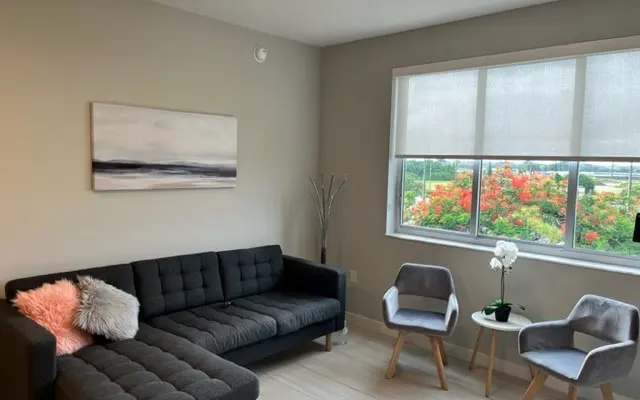 A modern living room featuring a dark gray sectional sofa with decorative pillows, two light gray chairs with wooden legs, and a small round table with a potted plant and an orchid. A large window shows a view of flowering trees outside.