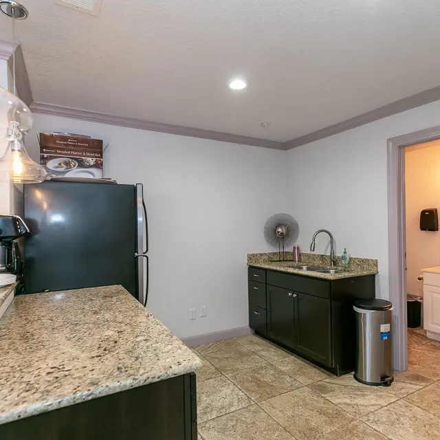 A modern kitchen featuring dark cabinetry and a granite countertop. A refrigerator, a coffee maker, and a sink are visible. The kitchen opens to a small hallway with a door leading to a bathroom.