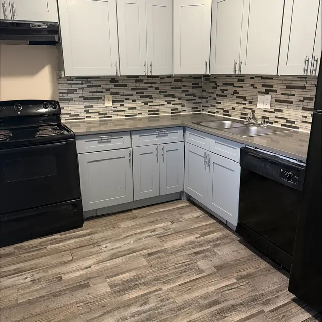 A modern kitchen featuring gray wooden cabinets, a black stove, a black dishwasher, and a tiled back wall with a mix of gray and white tiles. The floor is wooden-looking with a grayish tint.