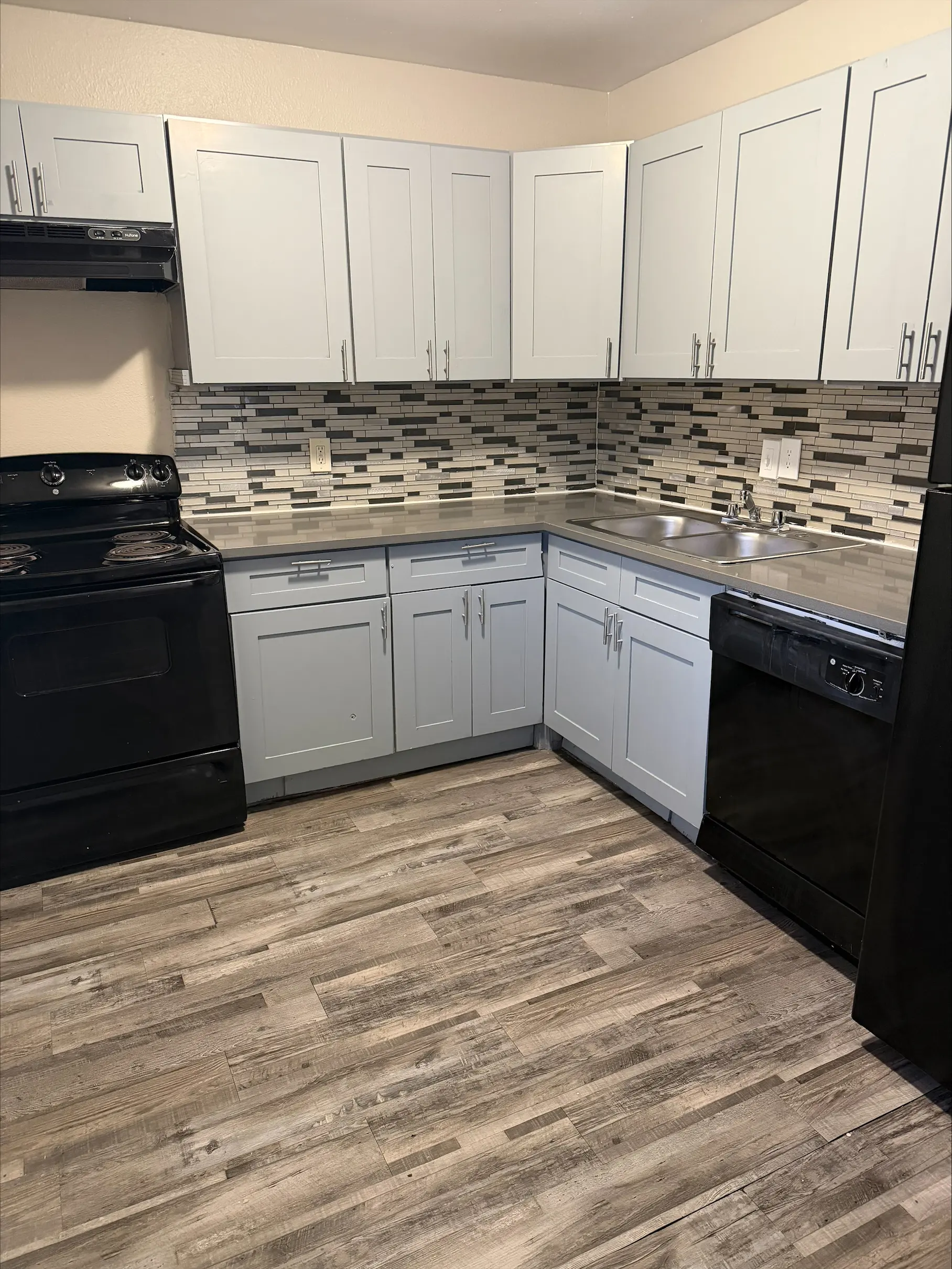 A modern kitchen featuring gray wooden cabinets, a black stove, a black dishwasher, and a tiled back wall with a mix of gray and white tiles. The floor is wooden-looking with a grayish tint.