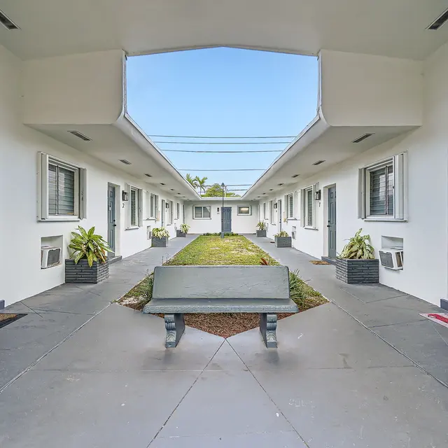 A view of a hotel-style courtyard with rooms on either side and a central grassy area. Each room has a dark door, and there are potted plants lining the walkway. A bench sits in the middle of the courtyard under a clear blue sky.