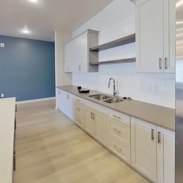 A spacious modern kitchen featuring white cabinets, a large countertop, and stainless steel appliances. The background has a contrasting blue wall, and there's a wooden cutting board with some food on the countertop.