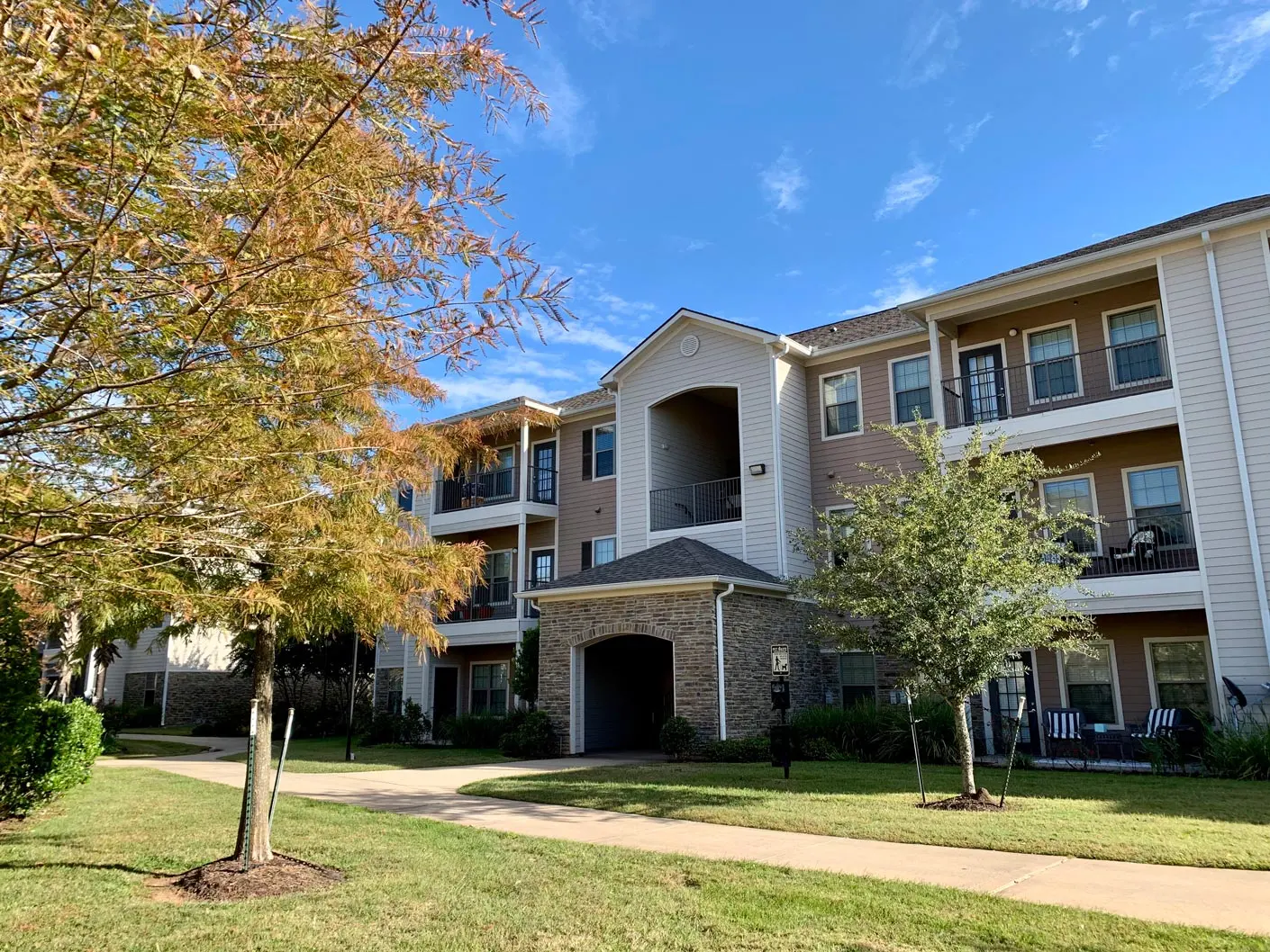 A sunny day view of a multi-story apartment building with trees and a pathway in front.