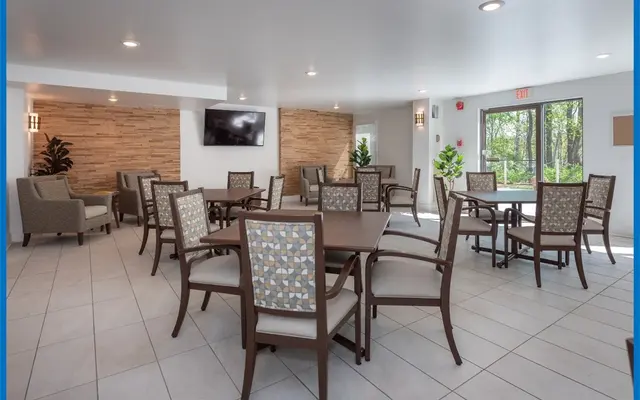 A bright and spacious dining area with several dining tables and chairs arranged neatly. The space has large windows allowing natural light, and there are plants in the corners. The walls are a combination of light colors and a textured wooden panel. There is a television mounted on the wall opposite the seating area.