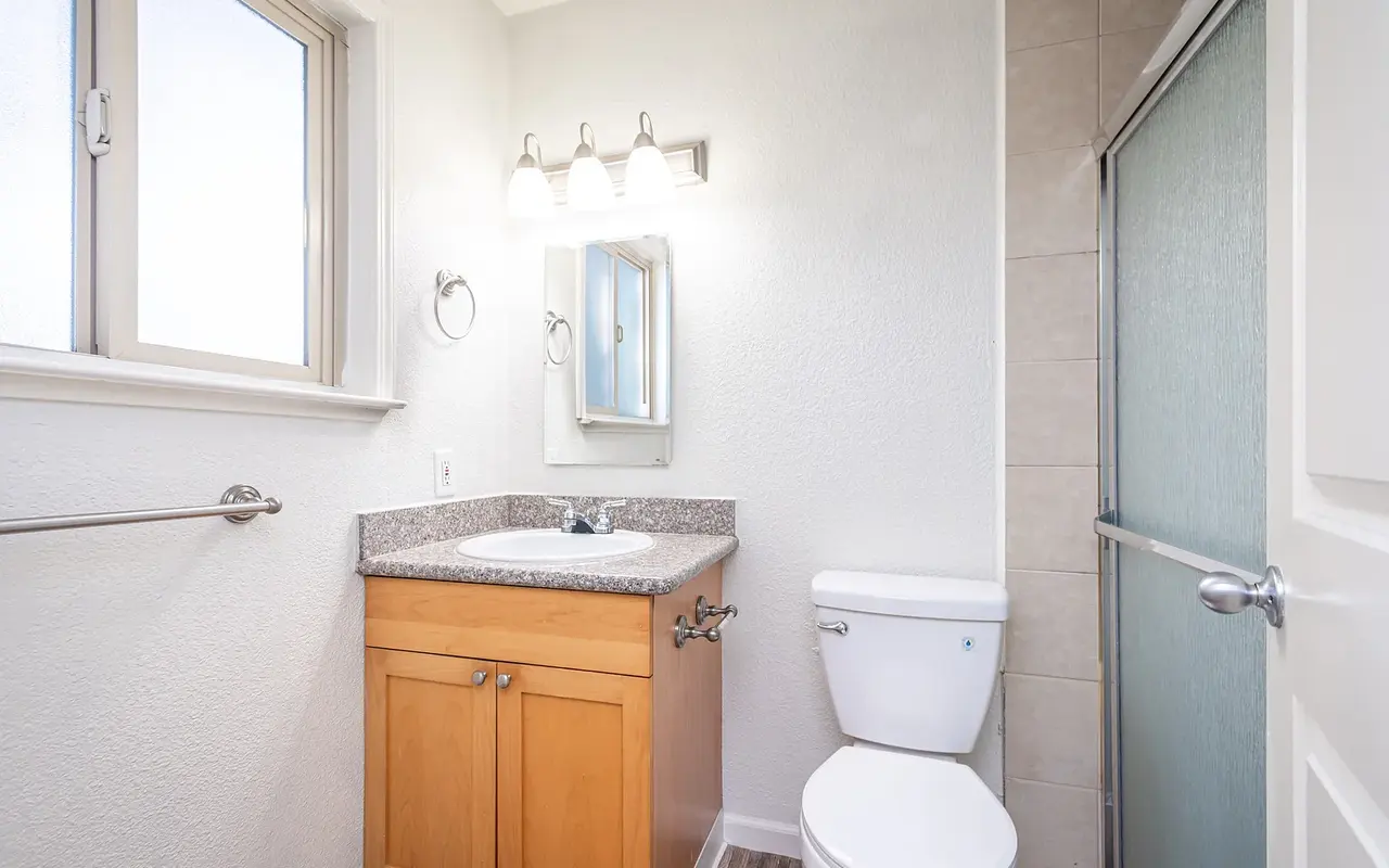 A modern bathroom featuring a wooden vanity with a granite countertop, a white toilet, and a glass-enclosed shower. Natural light enters through a large window, illuminating the space.