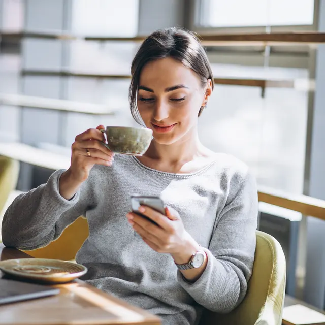A woman sitting in a cafe, smiling while holding a cup of coffee and looking at her phone. A laptop and a plate can be seen on the table.