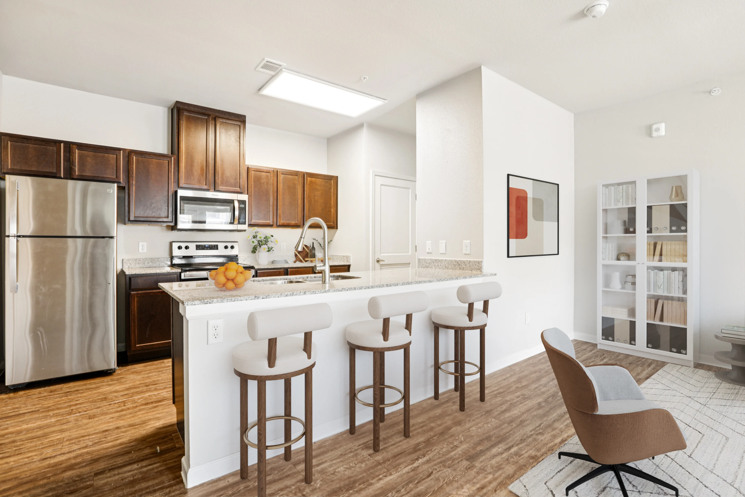 A modern kitchen featuring dark wooden cabinets, stainless steel appliances, and a large island with bar stools. The kitchen has a contemporary open-plan design leading to a cozy seating area with a chair and a bookshelf.