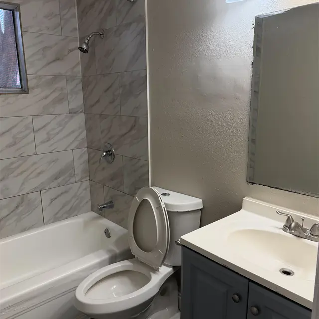 A modern bathroom with a bathtub, toilet, and sink. The walls are tiled in a light marble pattern and there is a small window.