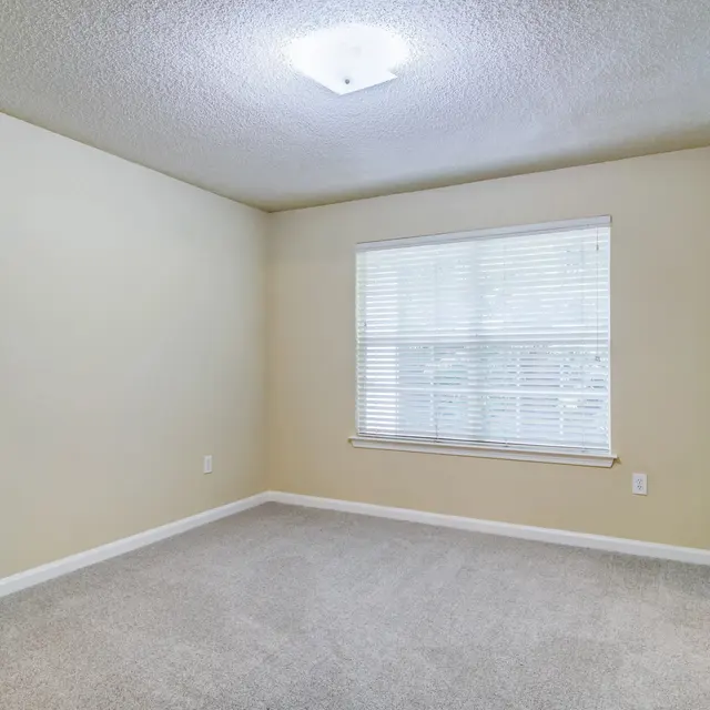 A spacious, empty room with beige walls and a window covered by white blinds. The floor is carpeted in light gray. There is a ceiling light fixture, and a door leading to another area on the right side.