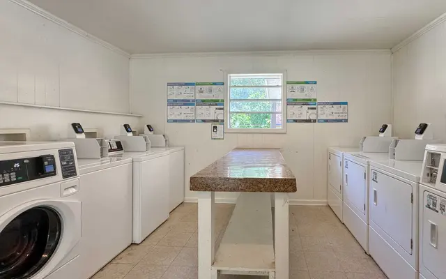A clean laundry room with several white washers and dryers, a countertop in the center, and informational posters on the walls.