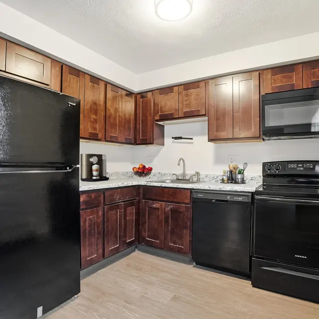 A compact kitchen with dark wood cabinetry, featuring a black refrigerator, stove, and microwave. The countertop includes a sink and kitchen accessories, with light-colored flooring.