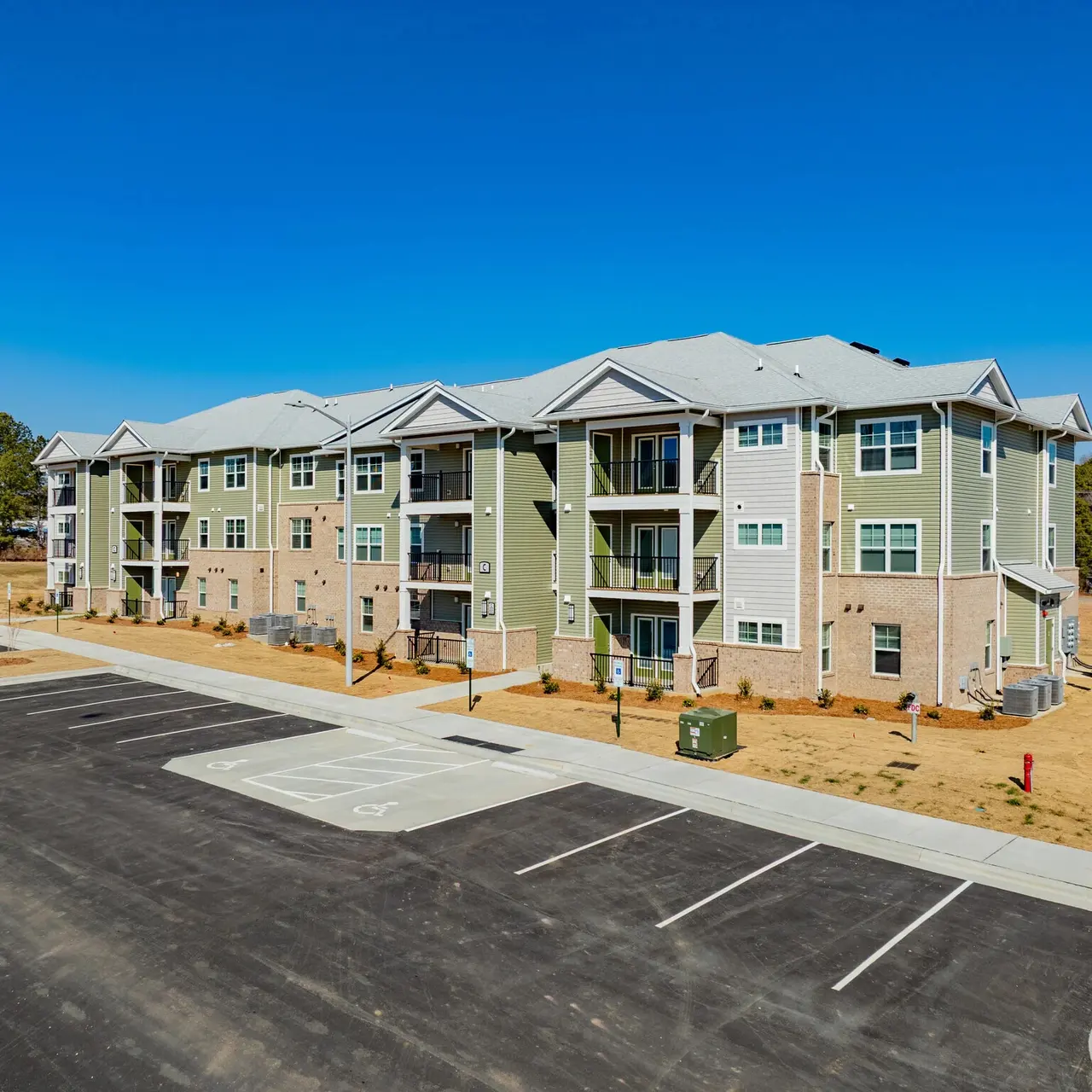 A newly constructed apartment complex with a modern design, featuring multiple exterior balconies and a landscaped area. A broad parking lot is in front with marked spaces, under a clear blue sky.