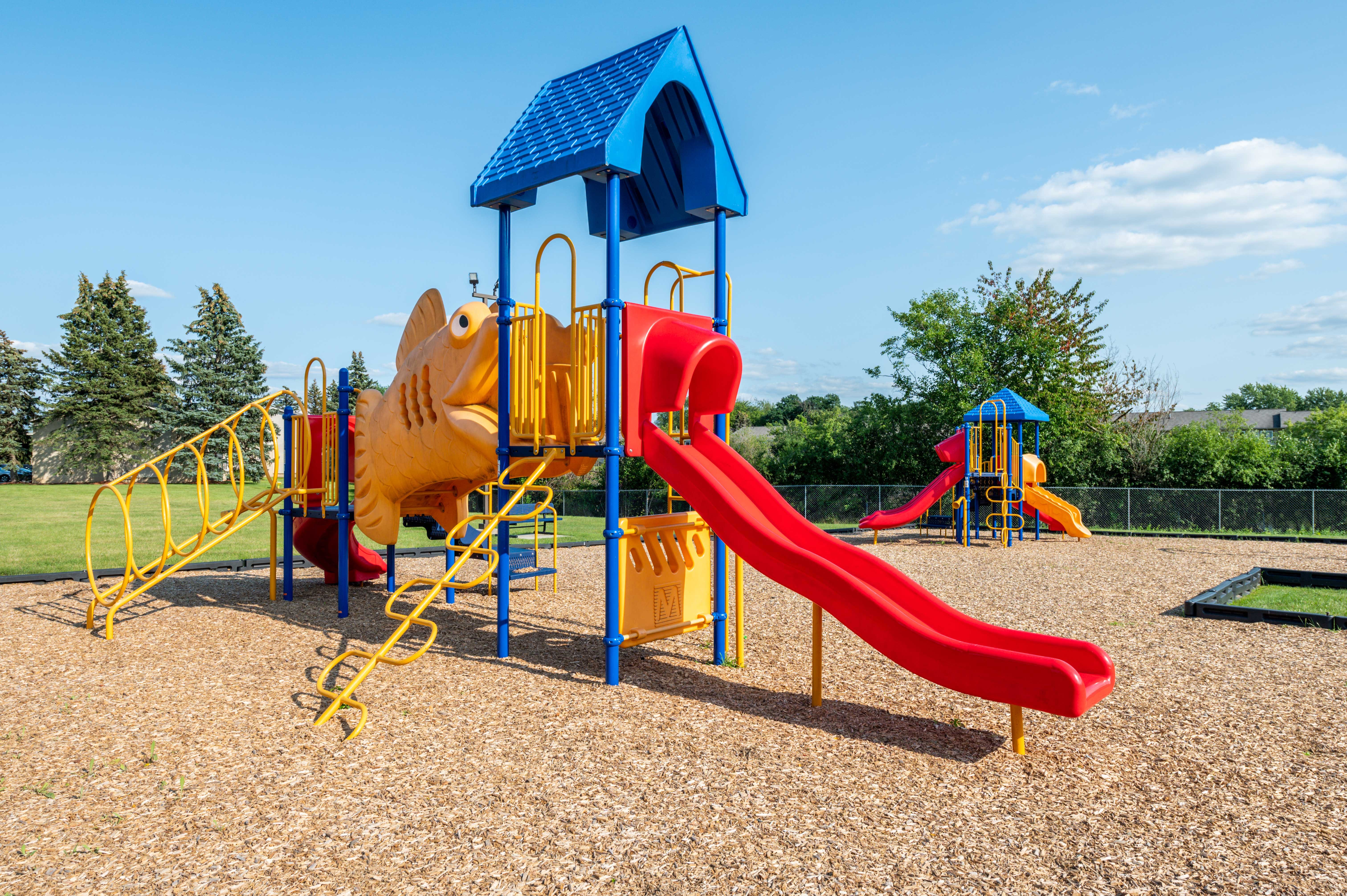 Colorful Fish-Themed Playground Colorful playground equipment featuring a large fish-shaped structure, slides, and climbing areas, set on a bed of wood chips under a clear blue sky.