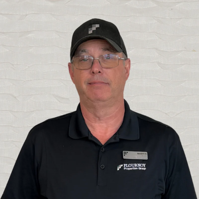 A middle-aged man wearing glasses, a black cap, and a black polo shirt with a name tag. He has short hair and is standing against a textured white background.