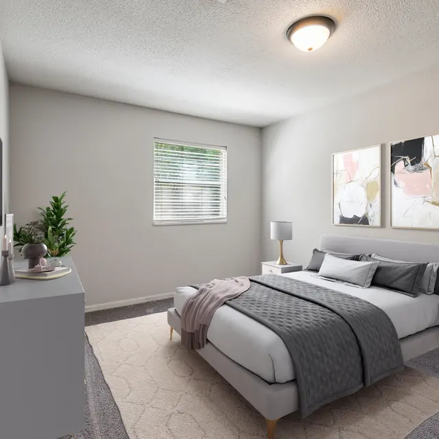 A modern bedroom featuring a bed with grey bedding, two bedside tables with lamps, artwork on the walls, and a small plant by a window.