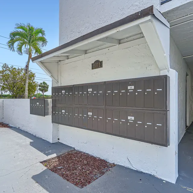 An exterior view of a mailbox area with multiple mailboxes under a roof. The setting includes a palm tree and a hallway leading to residential doors.