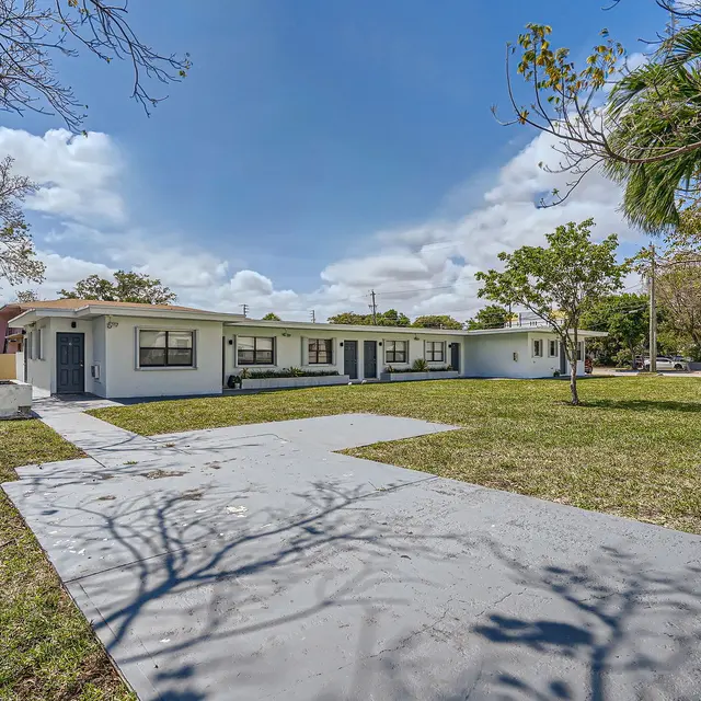 A single-story commercial building with a light-colored exterior, surrounded by green grass and trees. The building has multiple windows and an inviting entrance with a sidewalk leading up to it, set against a clear blue sky.