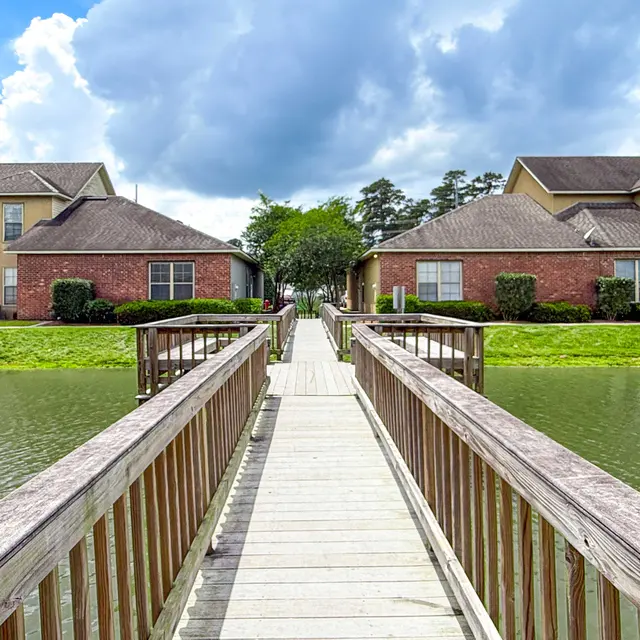 A wooden walkway leading between two residential buildings over a calm lake, surrounded by greenery and under a partly cloudy sky.