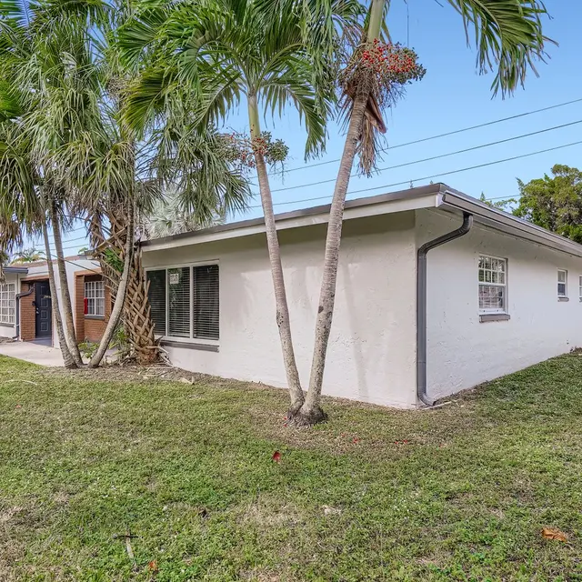 A view of a house from the backyard, featuring a white exterior and surrounded by palm trees and green grass.