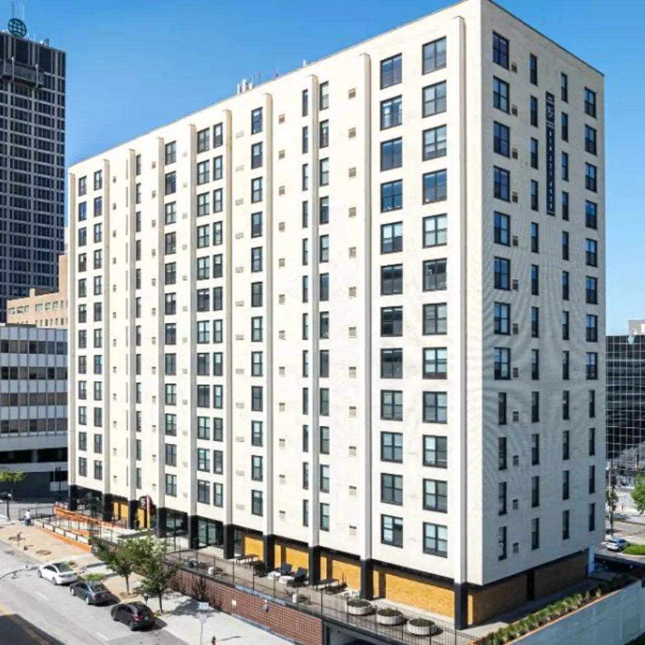 Modern Apartment Building in Urban Setting A modern seven-story apartment building with large windows, featuring a light-colored facade. The surrounding area has a mix of other buildings, including commercial and residential, with trees and parked cars nearby.
