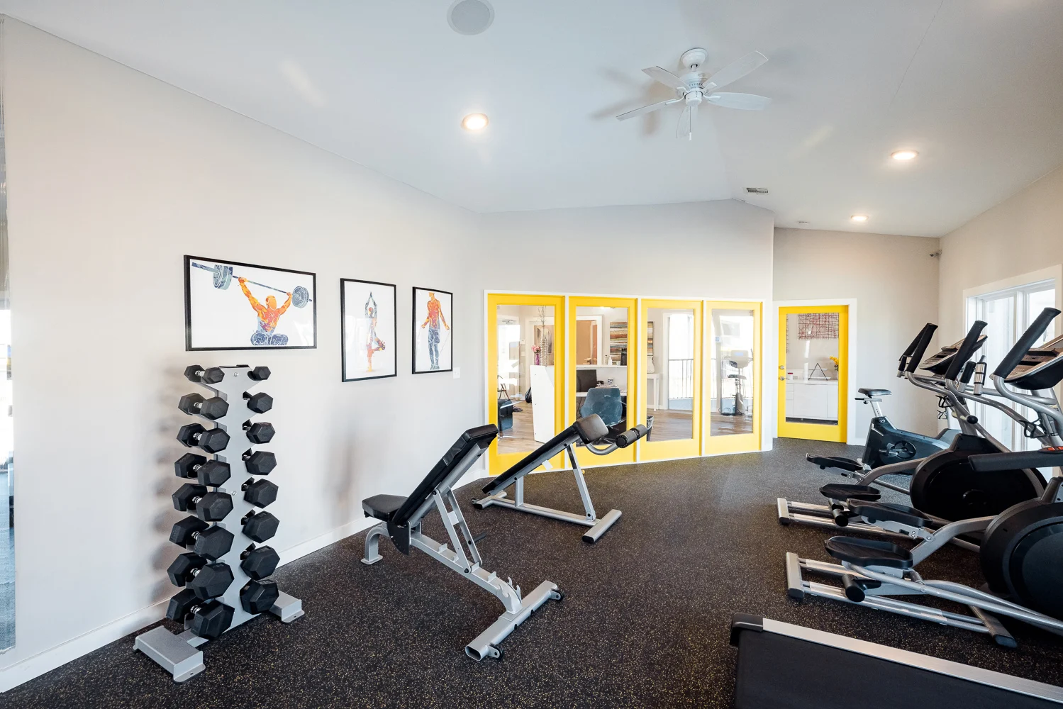 A modern gym interior featuring exercise equipment including a weight bench, dumbbells, and stationary bikes. The room has light-colored walls with framed art, a yellow accent wall, and large windows allowing natural light.