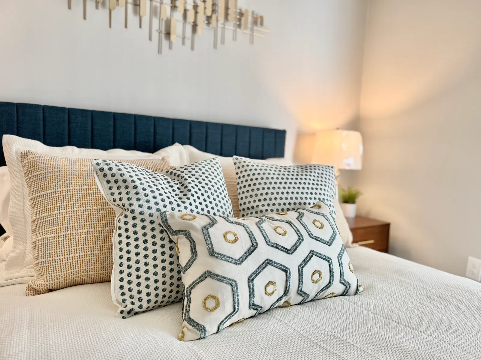A neatly made bed with decorative pillows in various patterns and a bedside lamp emitting a warm glow.