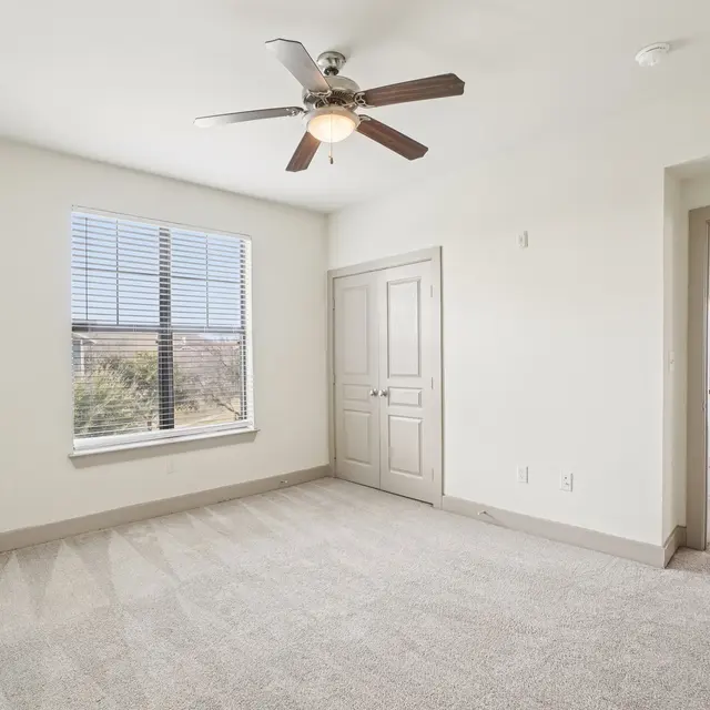 A spacious empty bedroom with a ceiling fan and large window letting in natural light. The walls are painted a light color, and the floor is carpeted. A door leads to another room in the background.