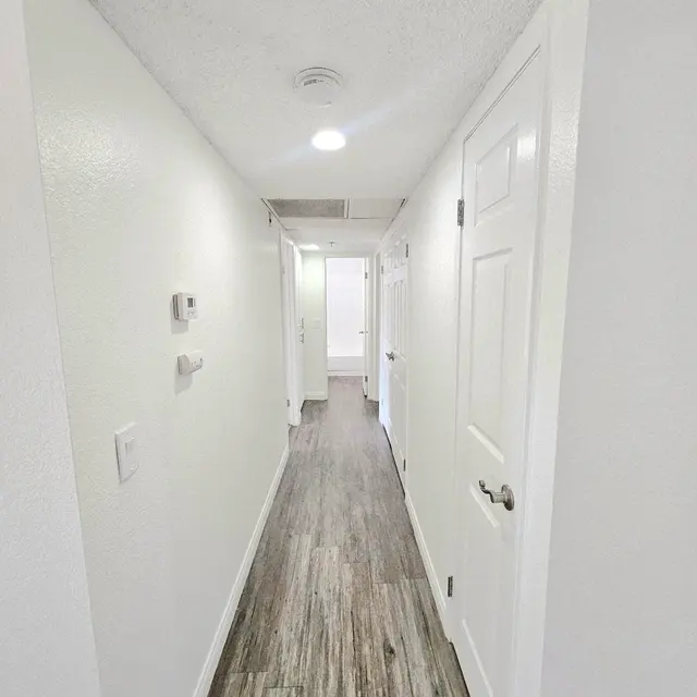 A long, narrow hallway with white walls and a wooden floor. It features two doors on the right side and one on the left side, leading to different rooms. Overhead lights illuminate the space, highlighting its clean and modern design.