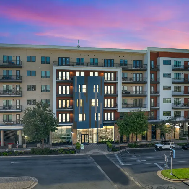 Modern apartment building with a mix of light and dark facades, featuring balconies and large windows under a vibrant sunset sky.