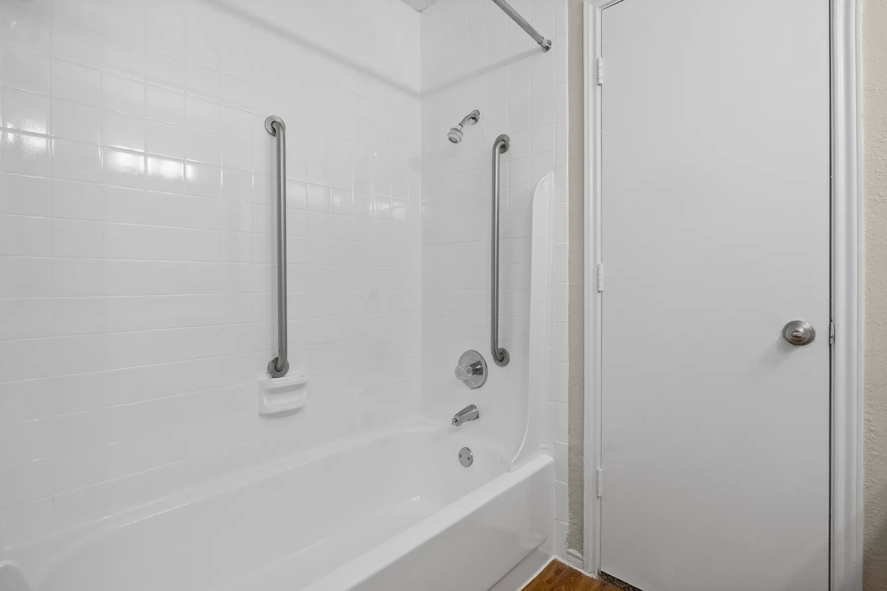 A clean and modern bathroom with a white bathtub, grab bars, and a door leading outside. The wall features white tiles and a single handle faucet.