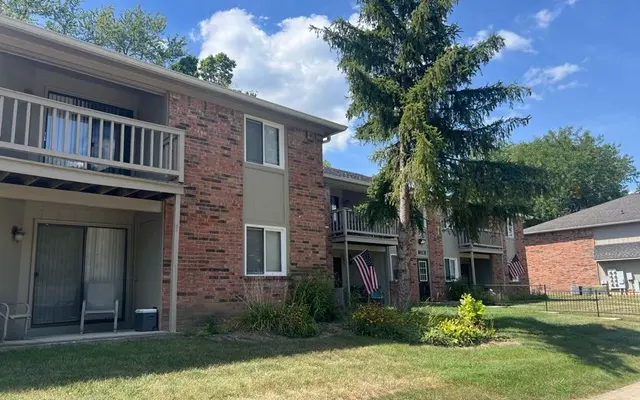 Apartment Complex View A street view of a multi-unit apartment complex with brick exterior, balconies, and a lush green lawn. There are trees and flags adorning the balconies.