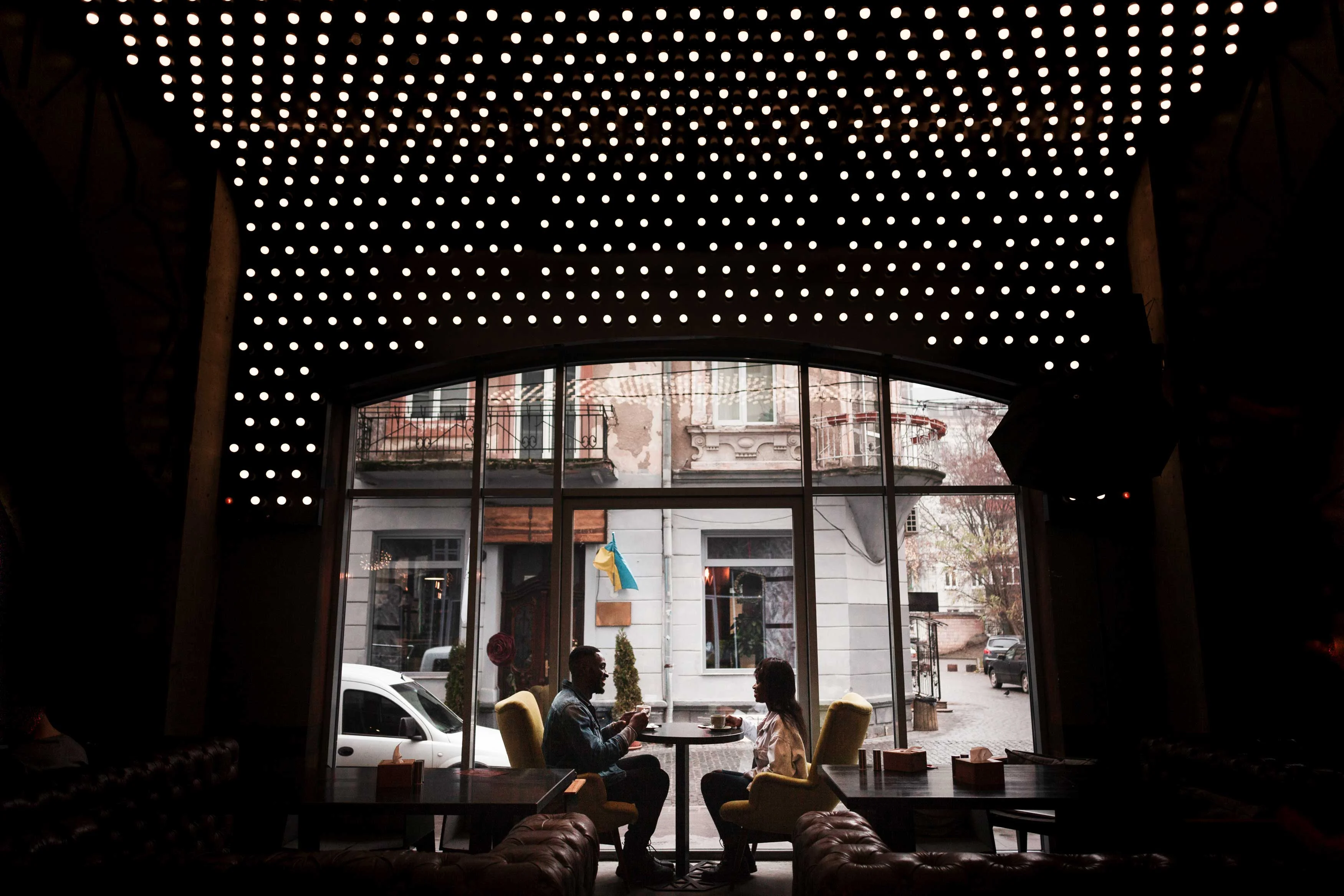 A Cozy Cafe Encounter Two people sitting at a table in a cafe, with a bright ceiling of lights and a window view of the street.