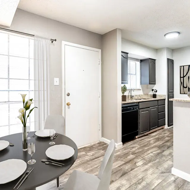 A modern dining area with a round black table and grey chairs, featuring place settings. The adjoining kitchen has dark cabinets and granite countertops.