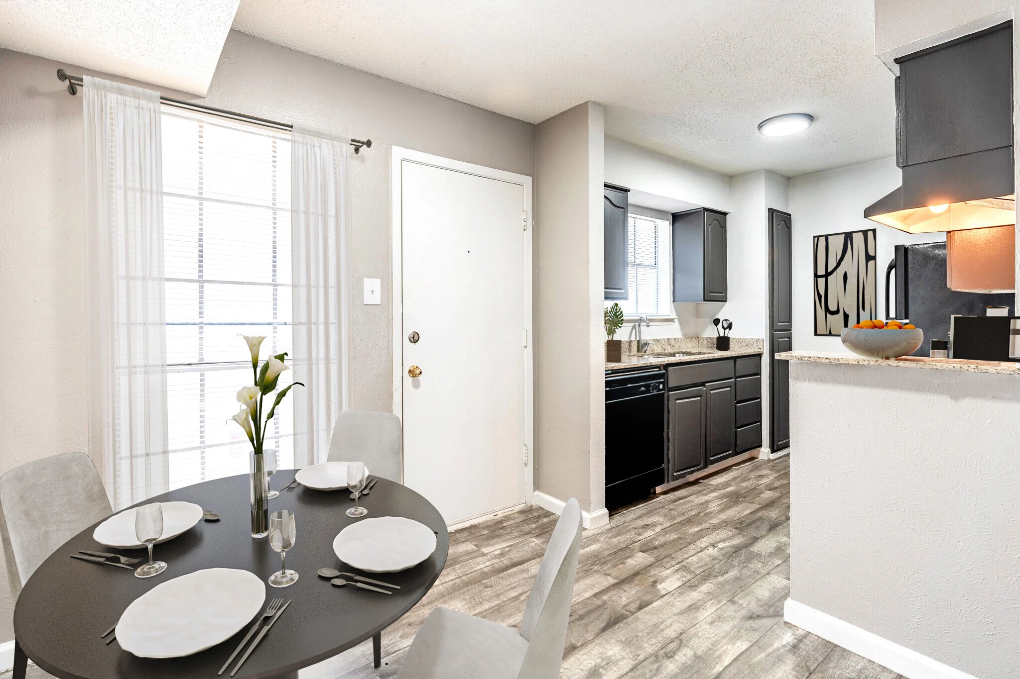 A modern dining area with a round black table and grey chairs, featuring place settings. The adjoining kitchen has dark cabinets and granite countertops.
