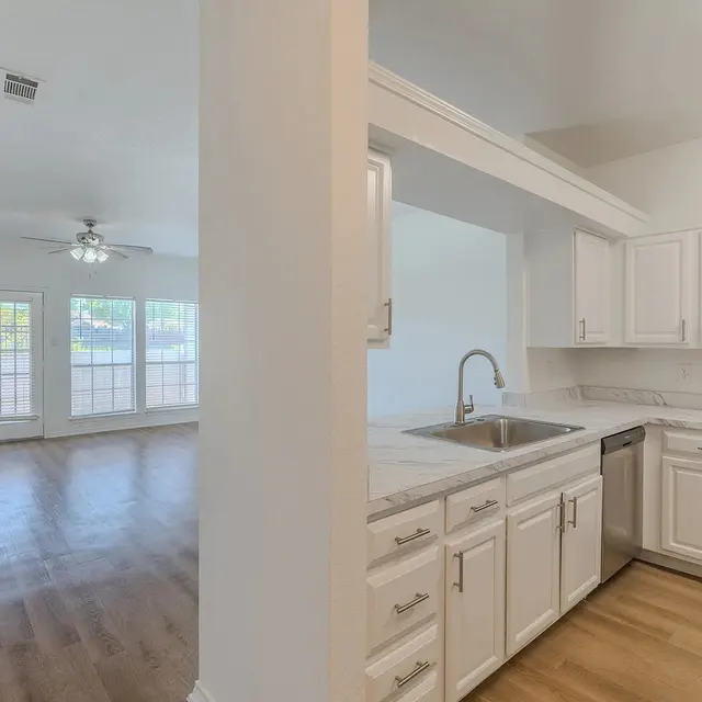 A modern kitchen with white cabinetry and stainless steel appliances that transitions into a spacious living room with large windows and a ceiling fan.