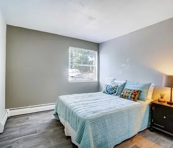 A cozy bedroom featuring a light blue quilted bedspread, decorative pillows, a bedside table with a lamp, and a window allowing natural light to enter.