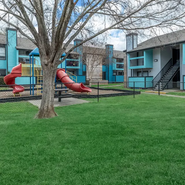 A playground with a red slide surrounded by green grass and a fenced area, next to two apartment buildings with blue and wooden exteriors.