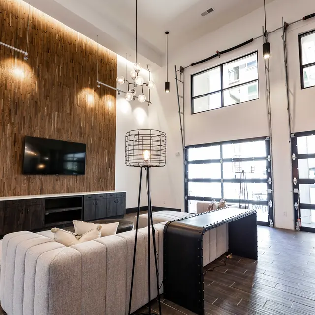 A spacious lobby area featuring a modern design with a beige sectional sofa, a black decorative table, and a wooden accent wall. The space is illuminated by pendant lights and has large glass doors.