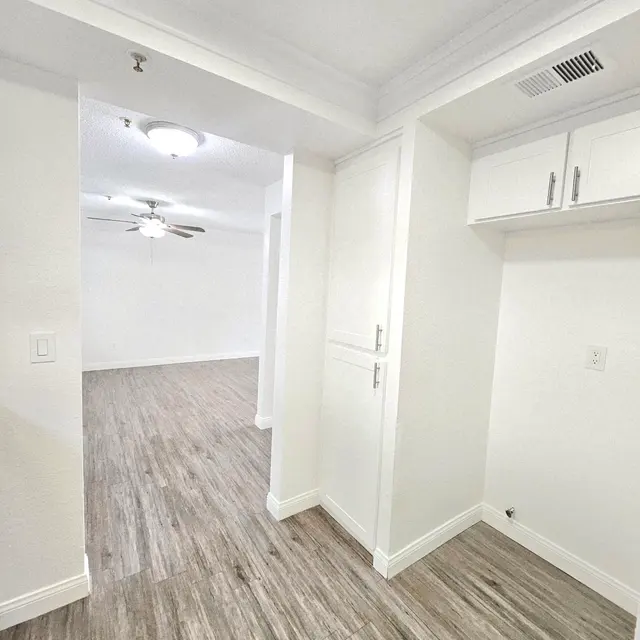 An interior view of a space with laminate flooring, looking through an open doorway into a well-lit area. The walls are painted white, and there is a ceiling fan visible.