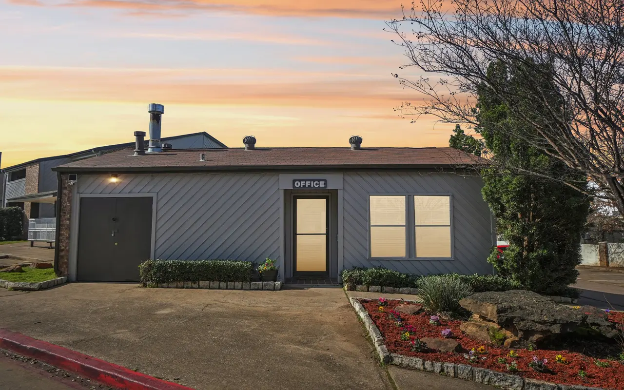 A single-story office building with a sign reading 'OFFICE' at the entrance. It features two large windows on the front and a door to the left. The surrounding area includes neatly trimmed hedges and colorful flower beds. The sky has a warm sunset glow.
