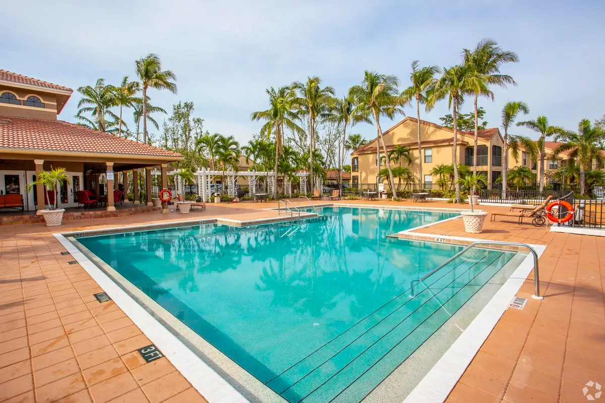 A clean and inviting swimming pool surrounded by palm trees and a well-kept patio area. Colorful buildings are visible in the background.