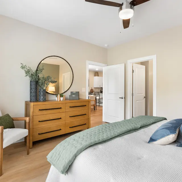 A bright and modern bedroom featuring a wooden dresser, a round mirror, and a comfortable bed with decorative pillows. There is a small armchair in the corner, and the room has natural light flowing in from the window.
