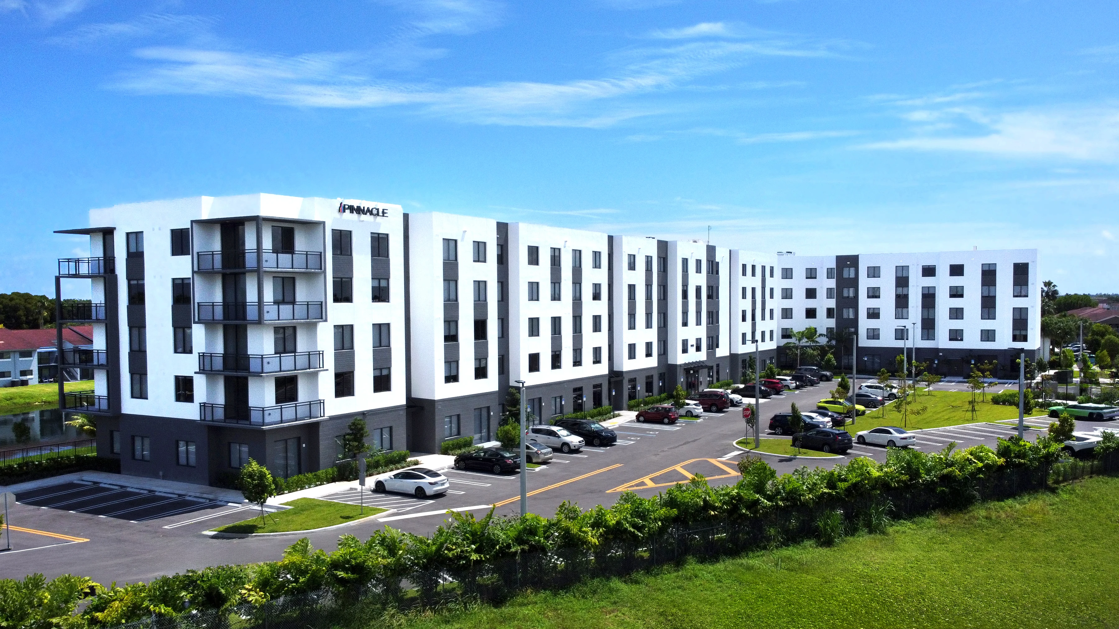 A modern apartment complex with three buildings, featuring a mix of white and dark facades, surrounded by a well-maintained lawn and parking spaces.