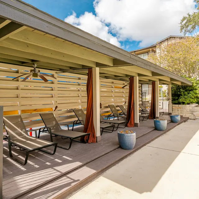 A poolside cabana with a wooden structure, featuring lounge chairs, a ceiling fan, and decorative pots. The area is shaded and surrounded by greenery, under a partly cloudy sky.