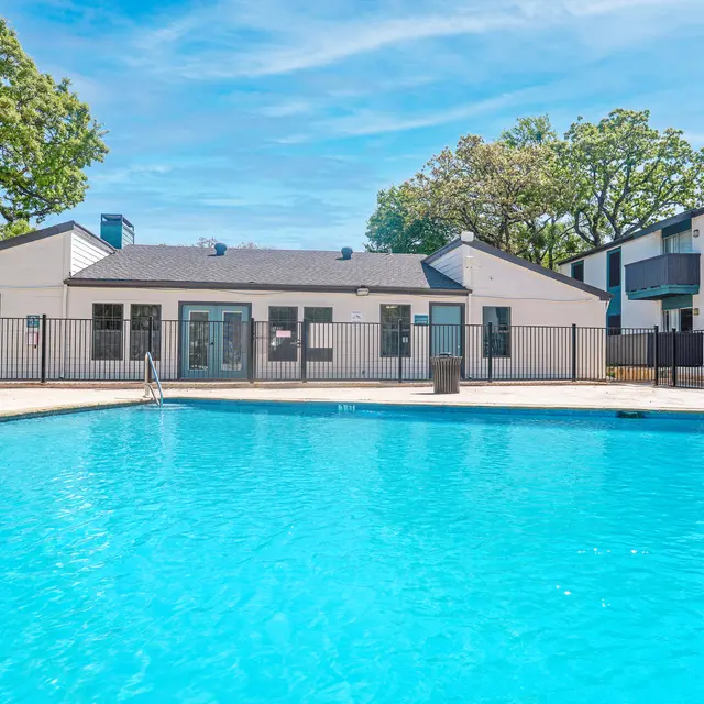 A bright and clear swimming pool surrounded by a fenced area, with buildings in the background and trees providing shade.