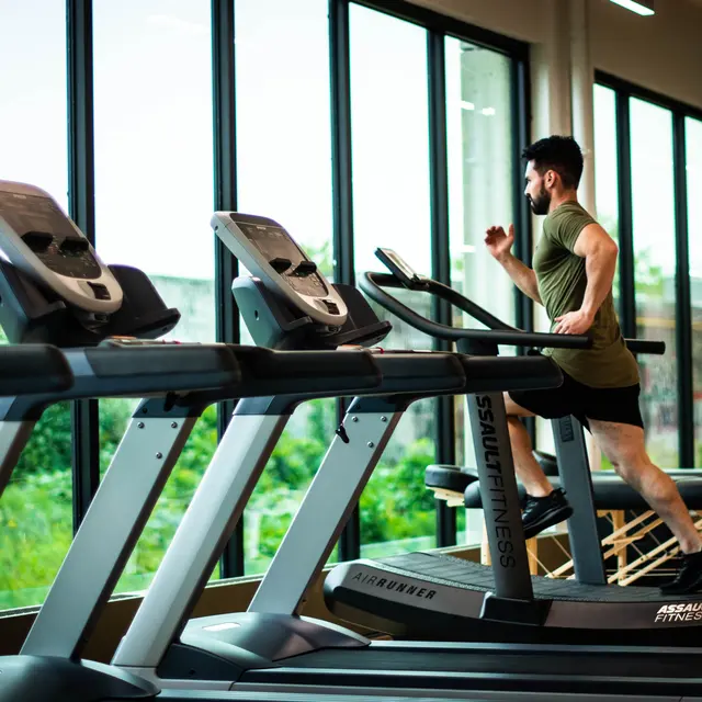 A man running on a treadmill in a modern gym with large windows showing greenery outside.